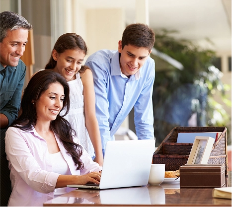 A group of colleagues gathers around a laptop on a desk, with one woman seated and typing while three others stand nearby, smiling and looking at the screen. The scene conveys teamwork and collaboration in a bright office setting.