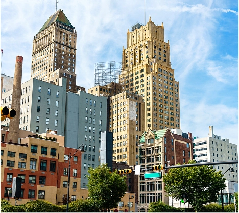 A cityscape featuring tall buildings with a clear blue sky. In the foreground, colorful, mid-rise structures line a street, with trees and a few smaller shops. The scene captures an urban downtown environment.